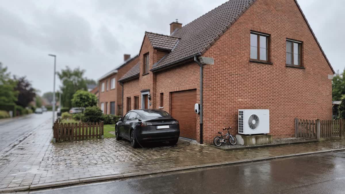 Een donkere elektrische wagen staat op de regenachtige kasseioprit van een Vlaamse bakstenen woning terwijl een laadkabel naar de wallbox naast de garagedeur loopt en een witte warmtepomp tegen de zijgevel staat met een kinderfiets ertussenin.
